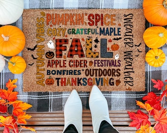 May include: A coir doormat with the word "FALL" in a plaid design, surrounded by autumn-themed words and images. Pumpkins and fall foliage frame the mat, with white boots in the foreground.