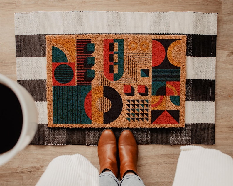 May include: Geometric patterned doormat with a black and white checkered rug underneath. The doormat features a design of colorful shapes including red, teal, orange, and black. Brown leather boots are visible at the bottom of the image, with light blue jeans and white ribbed fabric.