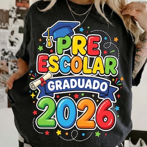 Peut inclure: T-shirt gris foncé avec un graphisme coloré célébrant la remise des diplômes de maternelle. Le motif comprend un chapeau de graduation, un diplôme et les mots "PRE ESCOLAR GRADUADO 2026" aux couleurs vives. Des étoiles et des confettis complètent le thème festif.
