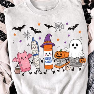 May include: A white crewneck sweatshirt featuring a colorful Halloween-themed graphic of cartoon school supplies including a pink eraser bat, a mummy pen, a glue stick in a witch hat, a stapler pumpkin, and a ghost calculator holding a pumpkin bucket.