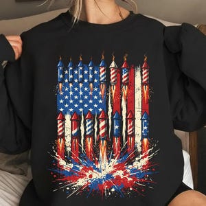 Könnte beinhalten: Schwarzes Sweatshirt mit einer Grafik der amerikanischen Flagge und Feuerwerk. Das Flaggen-Design enthält rote und weiße Streifen mit blauen Sternen, und das Feuerwerk wird als Raketen mit Flammen und Farbausbrüchen dargestellt.