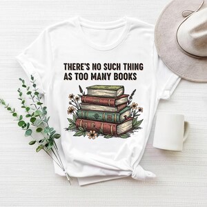 Peut inclure: T-shirt blanc avec une illustration d'une pile de livres, des fleurs et le texte "THERE'S NO SUCH THING AS TOO MANY BOOKS". Un chapeau et une tasse blanche sont également visibles.