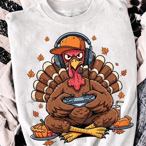 Puede incluir: Una camiseta blanca con el gráfico de un pavo marrón que lleva una gorra de béisbol naranja y auriculares azules con micrófono. El pavo sostiene un mando de videojuegos, rodeado de hojas de otoño naranjas, una tarta de calabaza y una bebida.