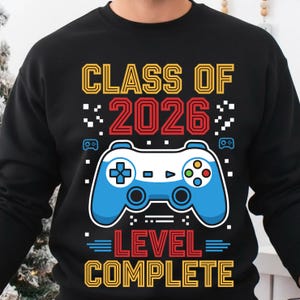 Peut inclure: Sweat-shirt noir avec une manette de jeu vidéo bleue et blanche. Le texte indique "CLASS OF 2026" en jaune et rouge, et "LEVEL COMPLETE" en rouge et jaune.