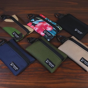 Könnte beinhalten: Eine Sammlung kleiner, rechteckiger Reißverschlusstaschen in verschiedenen Farben und Mustern. Designs umfassen Camouflage, Blumen, Schwarz, Marineblau, Olivgrün und Beige. Jede Tasche hat einen schwarzen Reißverschluss und ein Etikett mit "Doston MFG Made in USA".