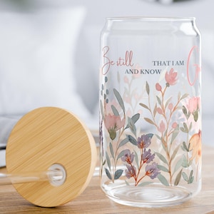 May include: A clear glass can-shaped drinking glass with a floral design and the words "Be still and know" in pink and black. The glass has a bamboo lid and a clear straw. The floral design includes pink, purple, and green flowers.