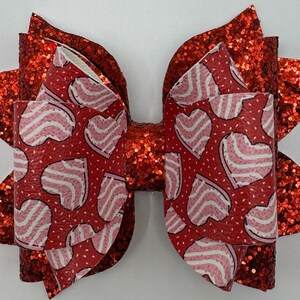May include: A red glitter hair bow with a white and pink heart pattern. The bow is made of fabric and has a layered design.