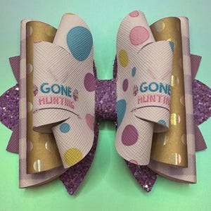 May include: A white bow with pink, blue, and yellow polka dots. The bow has a purple glitter center and gold glitter edges. The bow has the text "Gone Hunting" on both sides.