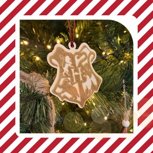 May include: A wooden Christmas tree ornament featuring the Hogwarts crest. The ornament is beige with a detailed design and a red string for hanging. It is set against a backdrop of a Christmas tree with lights.