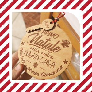 Può includere: Un ornamento natalizio rotondo in legno con una parte superiore a forma di chiave e un cordino rosso. L'ornamento presenta un testo inciso in italiano, tra cui "Primo Natale nella nostra NUOVA CASA" e i nomi Michele e Maria Giovanna, con l'anno 2024.