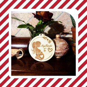 Può includere: Ornamento rotondo in legno chiaro con la silhouette di una donna incinta e il testo italiano "Aspettando Te" (Ti aspetto) e un cuore. L'ornamento è forato per essere appeso. Sullo sfondo un vaso con una rosa e altri oggetti decorativi.