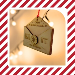 May include: A wooden Christmas decoration in the shape of an envelope, featuring the engraved text "CARO BABBO NATALE" and "LISTA DEI DESIDERI". The ornament includes a snowflake design and the word "EXPRESS". It is suspended by a red string.