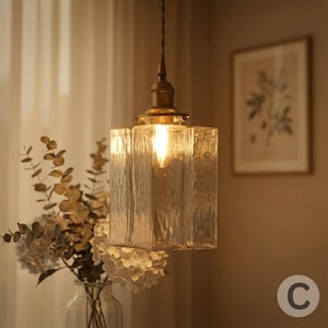 May include: A pendant light with a clear, textured glass shade and a brass-coloured metal base. The light fixture has a single bulb illuminating the textured glass. The light hangs from a cord, and is a decorative element.