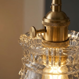 May include: Close-up of a vintage-style light fixture. The fixture features a clear, textured glass shade with a scalloped edge and a brass-colored metal socket. A small, round knob is visible on the side of the socket.