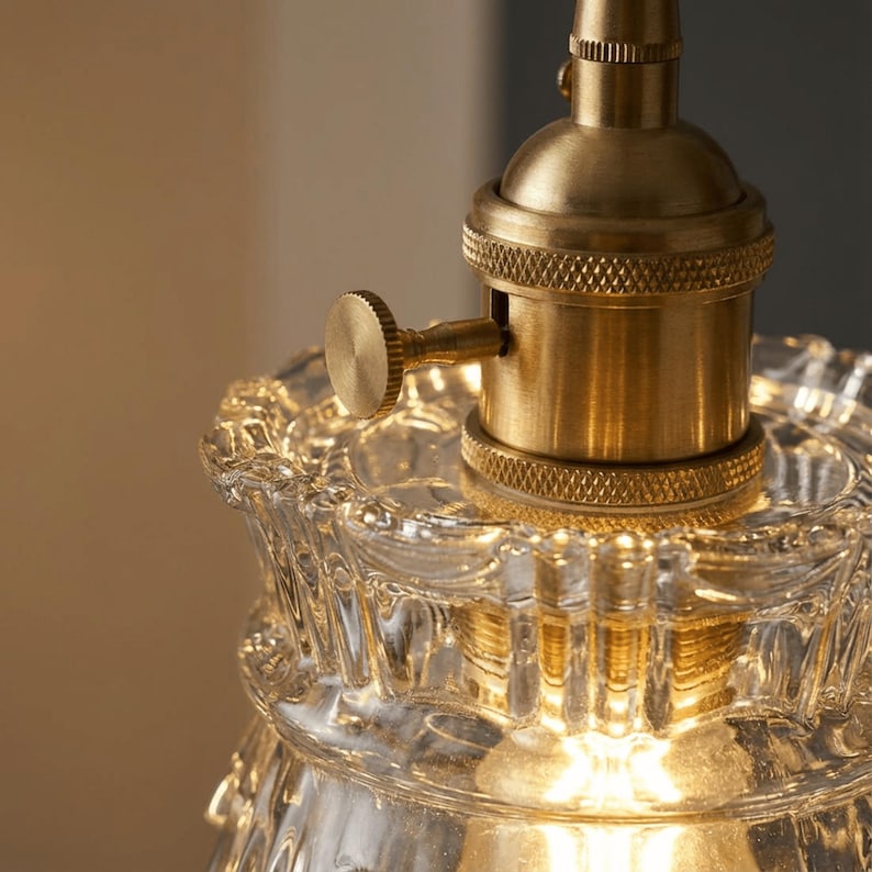 May include: Close-up of a vintage-style light fixture. The fixture features a clear, textured glass shade with a scalloped edge and a brass-colored metal socket. A small, round knob is visible on the side of the socket.