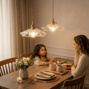 May include: Two pendant lights with clear, scalloped glass shades hang above a wooden dining table. The lights have brass-colored fixtures and are suspended by twisted cords. A woman and a child are seated at the table.