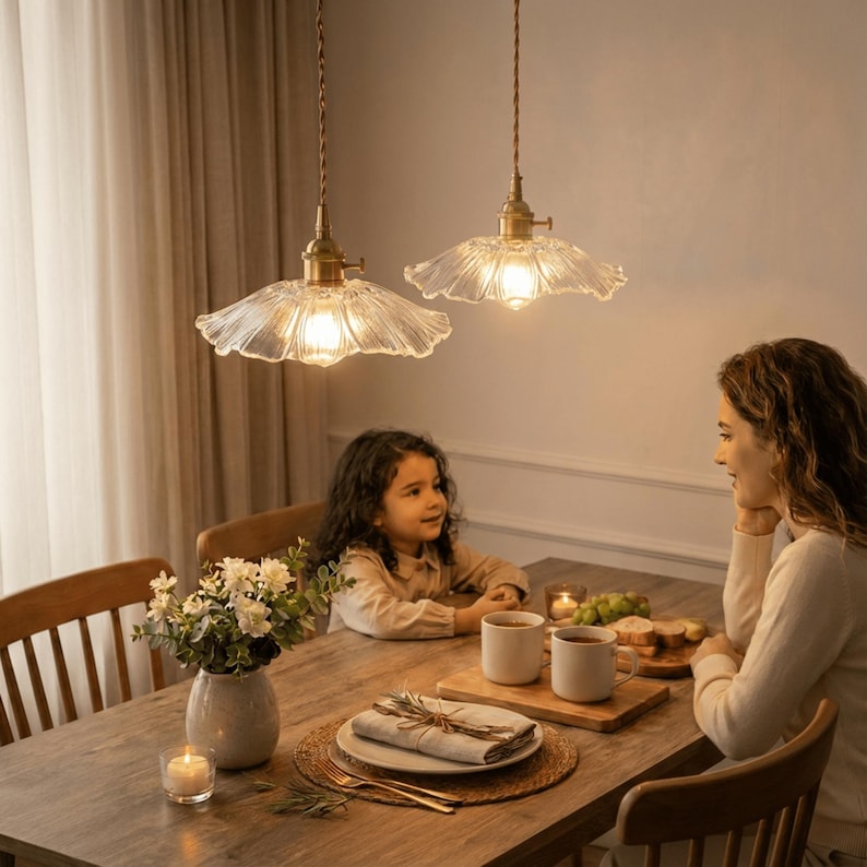 May include: Two pendant lights with clear, scalloped glass shades hang above a wooden dining table. The lights have brass-colored fixtures and are suspended by twisted cords. A woman and a child are seated at the table.