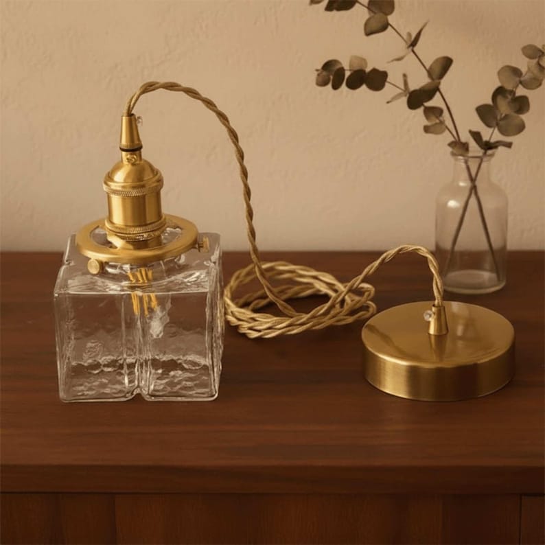 May include: A clear, square glass pendant light with a textured surface and brass-coloured fittings. It features a braided cord and a matching ceiling rose. A small glass vase with greenery is in the background.