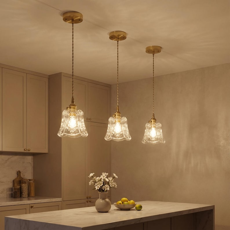 May include: Three pendant lights with clear, patterned glass shades and brass-colored hardware hang from the ceiling. The lights are suspended over a kitchen island with a marble countertop. The room has a neutral color palette.