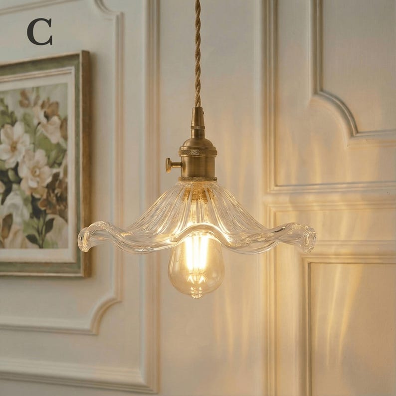 May include: A vintage-style pendant light with a clear, wavy glass shade and a brass-colored fixture. The light bulb is visible inside the shade. The light hangs from a twisted brown cord. A framed floral painting is in the background.