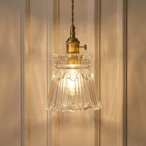 May include: A clear glass pendant light with a brass fixture and a ridged, bell-shaped shade. The light fixture hangs from a brown cord. The shade has a decorative, scalloped edge. The light bulb is visible inside the shade.