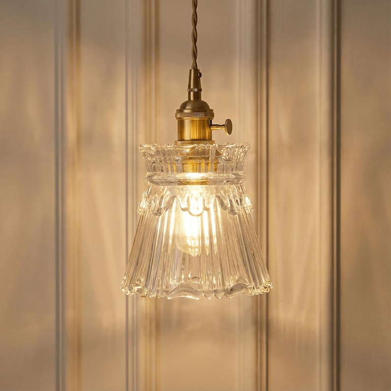 May include: A clear glass pendant light with a brass fixture and a ridged, bell-shaped shade. The light fixture hangs from a brown cord. The shade has a decorative, scalloped edge. The light bulb is visible inside the shade.