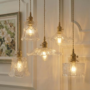 May include: Five vintage-style pendant lights with clear glass shades and brass fixtures hang from the ceiling. The shades have various designs, including floral patterns and textured surfaces, and are illuminated by warm-toned bulbs.