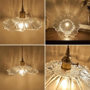 May include: Clear glass pendant light with a flower-shaped shade and brass hardware. The light fixture is suspended from a brown cord and emits a warm glow. The shade has a textured, rippled design.