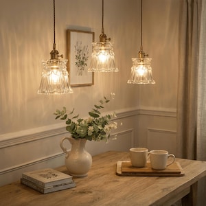 May include: Three pendant lights with clear glass shades and brass fixtures hang above a wooden table. The shades have a ruffled edge design. A white vase with greenery, two mugs, and a book are on the table.