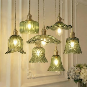 Op de afbeelding: Een verzameling van zes groene glazen hanglampen met messingkleurige hardware. Elke lamp heeft een uniek kapontwerp, waaronder bloemen en geschulpte randen. De lampen hangen aan het plafond.