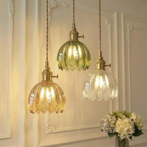 May include: Three pendant lights with scalloped glass shades in amber, green, and clear. Each light has a brass-coloured fixture and a decorative cord. The lights are hanging in front of a white wall, with a vase of flowers in the foreground.
