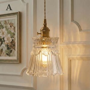 May include: A clear glass pendant light with a ruffled edge and a brass-colored fixture. The light bulb is visible inside the glass shade. The light hangs from a twisted cord.