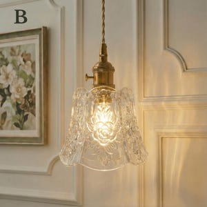 May include: A vintage-style pendant light with a clear, floral-patterned glass shade and brass hardware. The light fixture hangs from a twisted brown cord. The shade has a rose design in the center. The light is turned on, illuminating the glass.