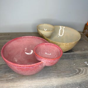 May include: Two ceramic serving bowls, each featuring a large main basin attached to a smaller side compartment. One bowl is finished in a speckled pink glaze, while the second bowl behind it is finished in a speckled beige and tan glaze.