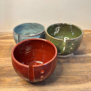 May include: Three ceramic yarn bowls in red, green, and blue. Each bowl features a smooth, glossy finish, a vertical slit for yarn, and three small circular holes on the side for guiding thread. The bowls are arranged in a cluster.