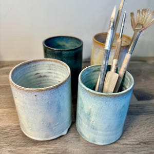May include: Three cylindrical ceramic cups with a glazed finish in shades of light blue, dark teal, and warm amber. The cups feature a smooth, slightly textured surface with visible rim detailing. Several artist paintbrushes are stored inside.