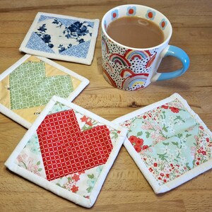 May include: A mug of tea sits beside four handmade fabric coasters. The coasters feature floral and geometric patterns in blue, green, yellow, and red. The mug has a blue handle and a rainbow design.