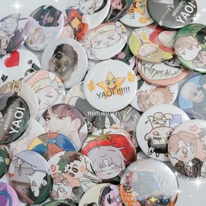 Puede incluir: Una colección de botones redondos y coloridos con varias ilustraciones y textos de estilo anime. Los botones presentan imágenes de personajes, el texto "Yaoi" y otros diseños. Los botones están dispersos sobre una superficie blanca.