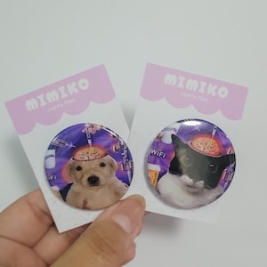 Puede incluir: Dos insignias redondas con ilustraciones de animales. Una muestra un golden retriever con un cerebro, la otra un gato blanco y negro con un cerebro. Ambas insignias tienen el texto "MIMIKO" en la tarjeta de respaldo.