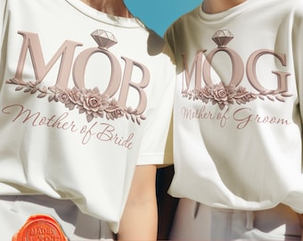 May include: Two white t-shirts with the text "MOB Mother of Bride" and "MOG Mother of Groom" in pink lettering with a diamond ring graphic and floral accents.