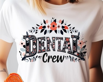 Op de afbeelding: Wit t-shirt met de tekst "DENTAL Crew" in een bloemenontwerp. Het woord "DENTAL" is in een decoratief lettertype, met roze en koraalroze bloemen en groene bladeren.