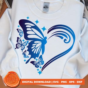 Könnte beinhalten: Ein weißer Pullover mit einem blauen und violetten Schmetterlingsdesign. Der Schmetterling hat die Form eines Herzens mit kleineren Schmetterlingen, die ihn umgeben. Der Text "MADE HEART STUDIO" befindet sich oben auf dem Pullover.