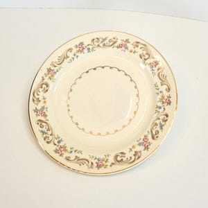 Può includere: Un piatto in ceramica bianca con un bordo dorato e un motivo floreale. Il piatto ha un bordo ondulato e un disegno dorato al centro.
