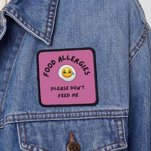 Puede incluir: Un parche cuadrado rosa con el texto "Food Allergies" en un círculo negro. Dentro del círculo hay un huevo de dibujos animados amarillo con una cara triste y una sola lágrima. Debajo del círculo está el texto "Please don't feed me".