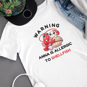 Può includere: Maglietta bianca con testo nero che recita "WARNING ANNA IS ALLERGIC TO SHELLFISH". Accanto al testo è raffigurata una langosta e una vongola di cartone animato.