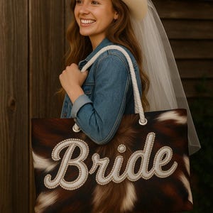 May include: A white cowhide tote bag with a white rope handle and the word "Bride" in white script with pearl accents. The bag is being held by a person wearing a white cowboy hat and a white veil.