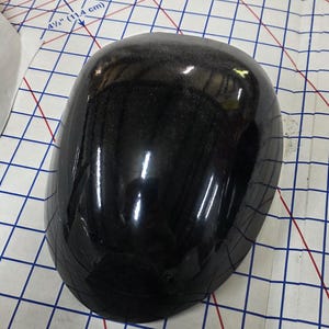 Pu&ograve; includere: Un oggetto nero lucido e arrotondato con una superficie maculata. L'oggetto &egrave; posizionato su una superficie a griglia con misure in pollici e centimetri.
