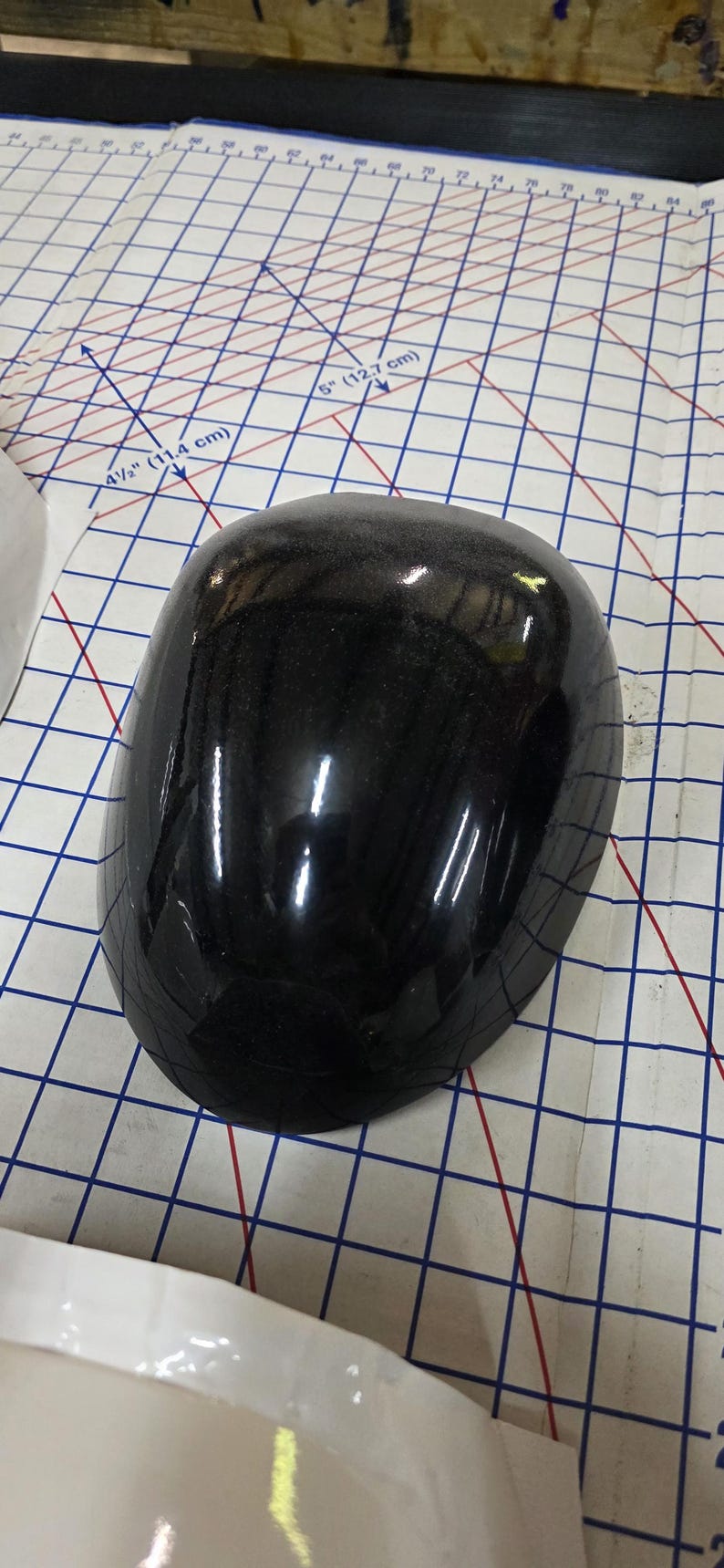 Pu&ograve; includere: Un oggetto nero lucido e arrotondato con una superficie maculata. L'oggetto &egrave; posizionato su una superficie a griglia con misure in pollici e centimetri.