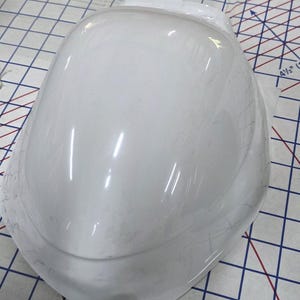 Könnte beinhalten: Ein weißes, glänzendes, helmformiges Objekt, möglicherweise ein Bauteil oder eine Form, liegt auf einer karierten Oberfläche. Das Objekt hat eine glatte, gebogene Oberfläche mit mehreren Lichtreflexen.