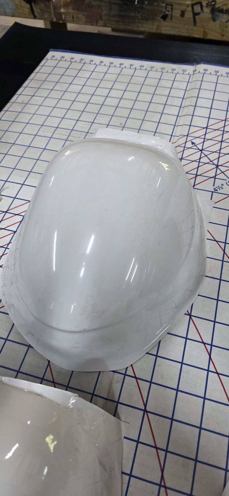 Pu&ograve; includere: Un oggetto bianco lucido a forma di casco, possibilmente un componente o uno stampo, &egrave; posto su una superficie a griglia. L'oggetto ha una superficie liscia e curva con diversi riflessi di luce.